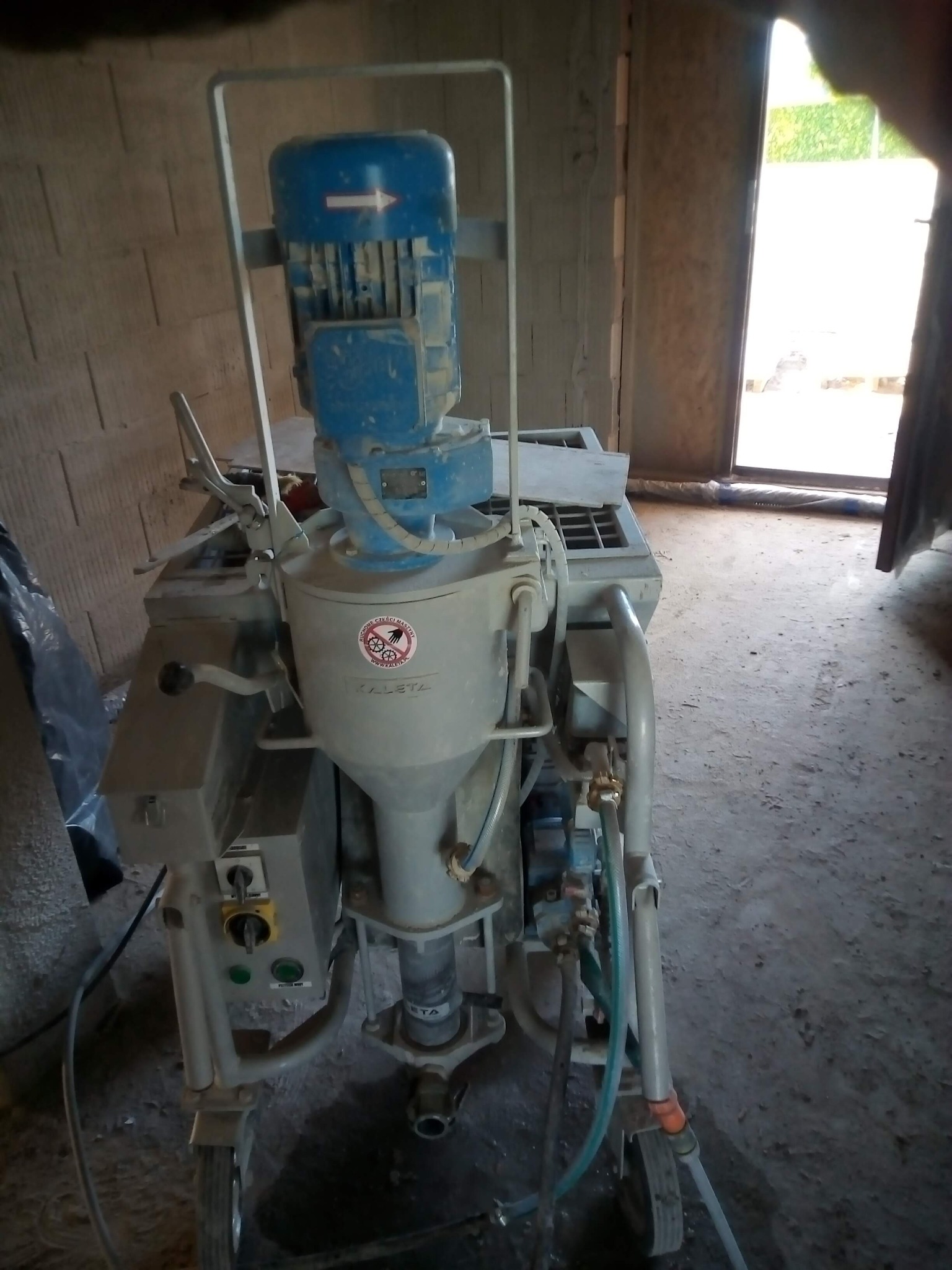 Maszyna do tynkowania KALETA z niebieskim silnikiem i naklejką ostrzegawczą, stojąca w surowym wnętrzu, prawdopodobnie przed tynkowaniem.