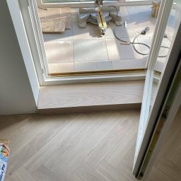 Eryk Czapla - Układanie jasnych paneli podłogowych w jodełkę francuską, widoczny próg drzwiowy i worek z klejem. W tle narzędzia do cięcia paneli.