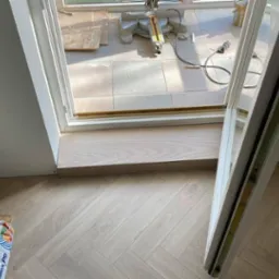 Układanie jasnych paneli podłogowych w jodełkę francuską, widoczny próg drzwiowy i worek z klejem. W tle narzędzia do cięcia paneli.