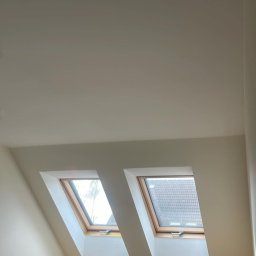 Eryk Czapla - Pokój na poddaszu z dwoma oknami dachowymi, biurkiem i krzesłem. Ściany pomalowane na jasny kolor, widoczne grzejniki. Ujęcie z dołu, podkreślające skos dachu.