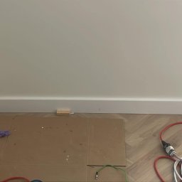 Eryk Czapla - Montaż białych listew przypodłogowych w Krakowie. Ułożona podłoga w jodełkę, zabezpieczone narożniki taśmą malarską. Widoczne narzędzia pracy.