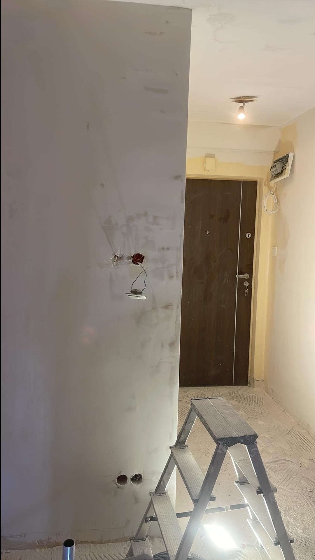 Ściana po szpachlowaniu z widocznymi puszkami elektrycznymi, drabina w tle. Prace wykończeniowe w korytarzu z brązowymi drzwiami wejściowymi.