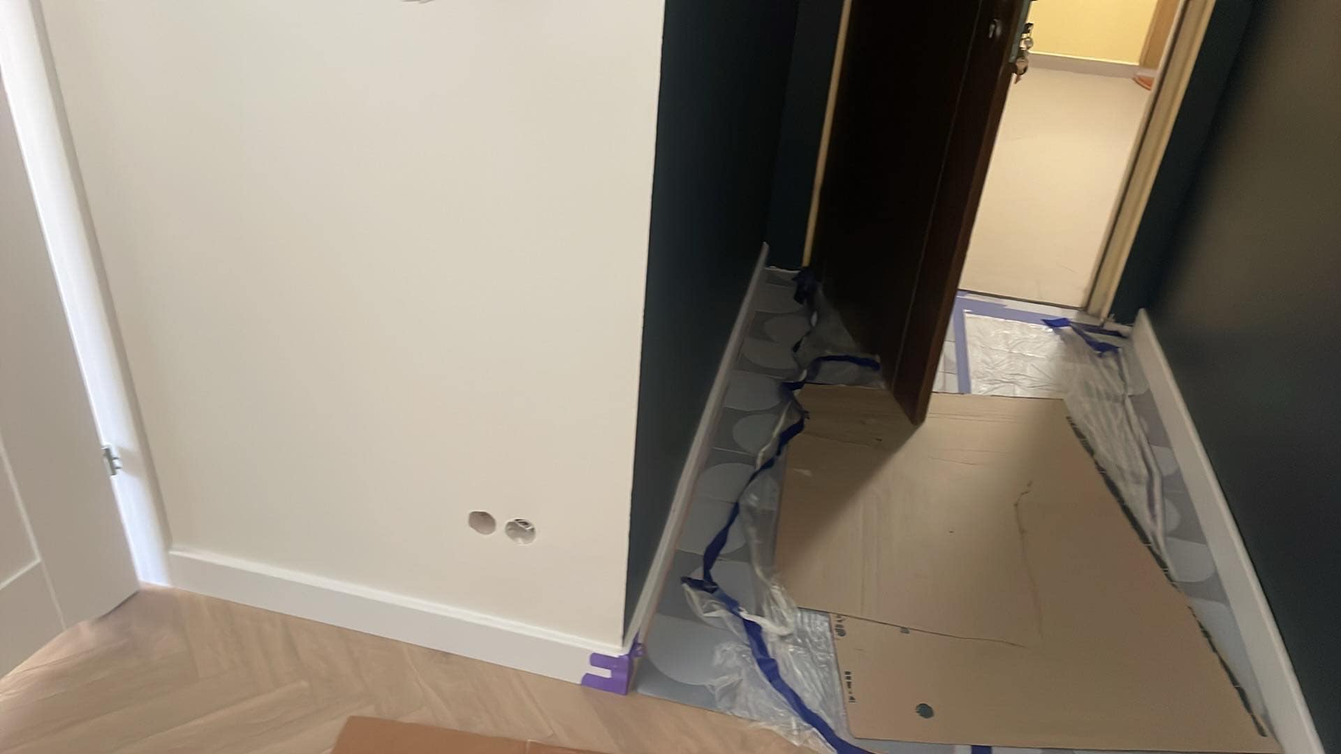 Przygotowanie do prac: zabezpieczona folią i kartonem podłoga w jodełkę, ściany w kolorach bieli i grafitu, widoczne gniazdka elektryczne i listwy przypodłogowe.