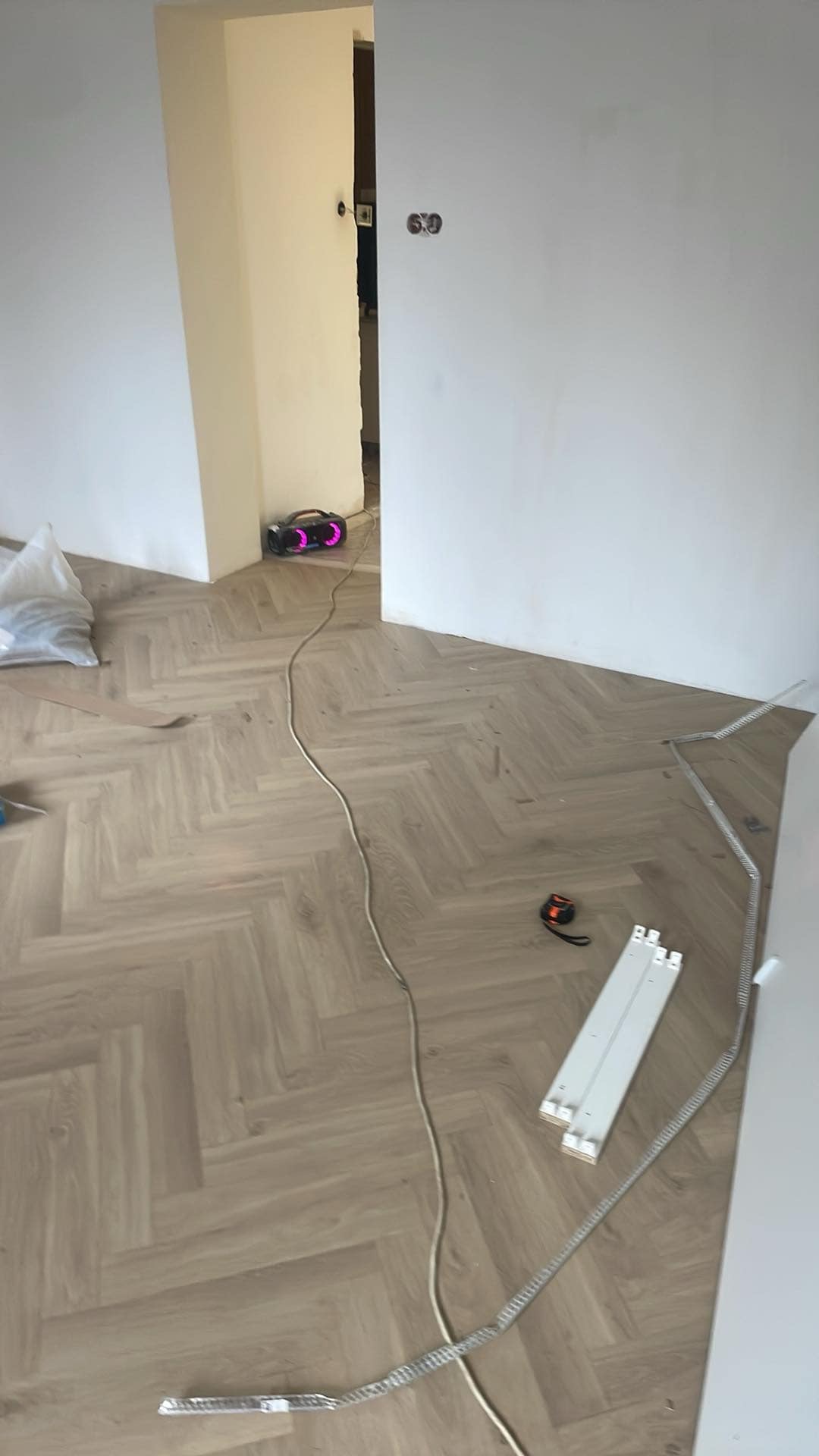 Ułożona podłoga w jodełkę, widoczne listwy przypodłogowe, taśma miernicza i głośnik w tle. Parkiet w odcieniach beżu, świeżo po montażu.