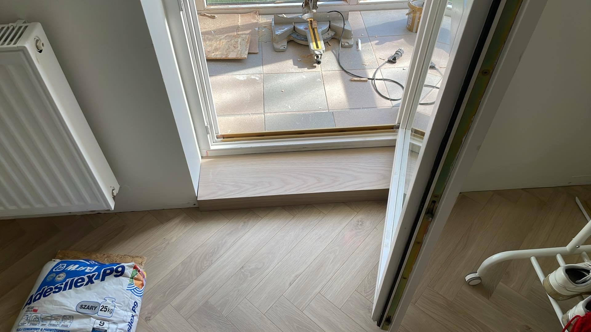 Układanie jasnych paneli podłogowych w jodełkę francuską, widoczny próg drzwiowy i worek z klejem. W tle narzędzia do cięcia paneli.