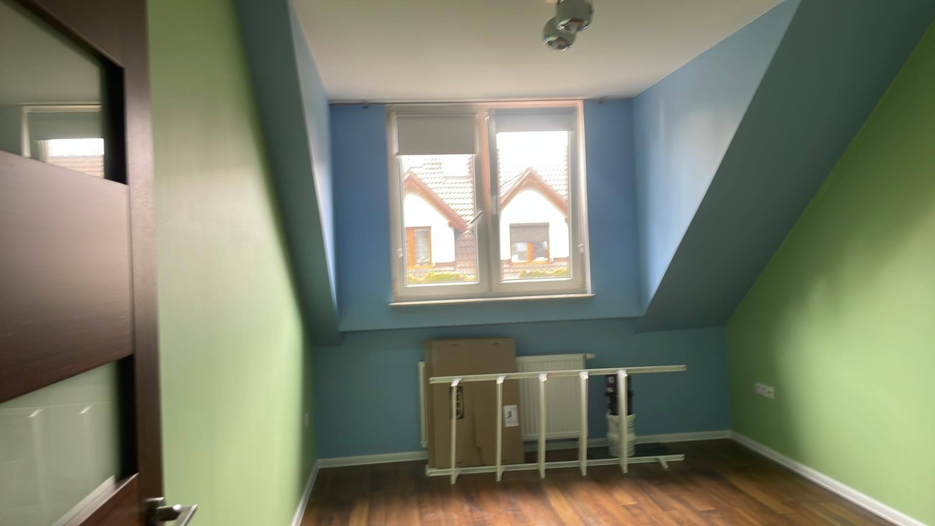 Poddasze z oknem i skośnymi ścianami, pomalowane na zielono i niebiesko. Widoczne panele podłogowe, grzejnik i kartonowe pudło. Przygotowanie do prac wykończeniowych.