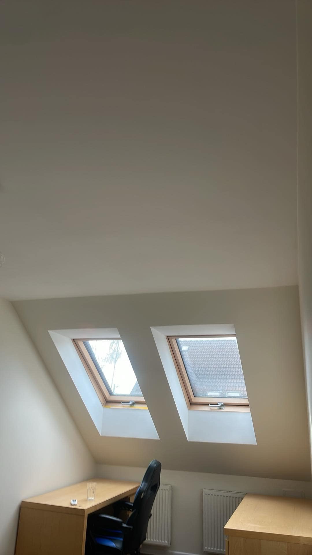 Pokój na poddaszu z dwoma oknami dachowymi, biurkiem i krzesłem. Ściany pomalowane na jasny kolor, widoczne grzejniki. Ujęcie z dołu, podkreślające skos dachu.