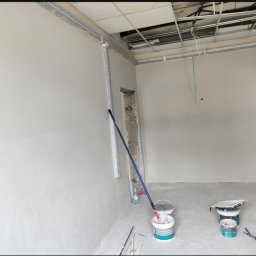 Move- LINE - Wnętrze pokoju w trakcie prac wykończeniowych; świeżo pomalowane ściany na biało, widoczne narzędzia malarskie i puszki z farbą. Sufit z instalacją.