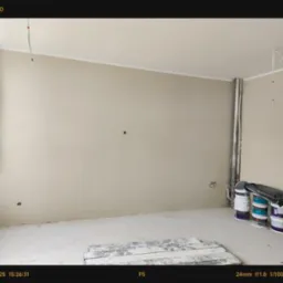 Pokój w trakcie malowania: ściany w beżowym odcieniu, widoczne puszki z farbą, przygotowane do dalszych prac wykończeniowych. Minimalizm i stonowana kolorystyka.