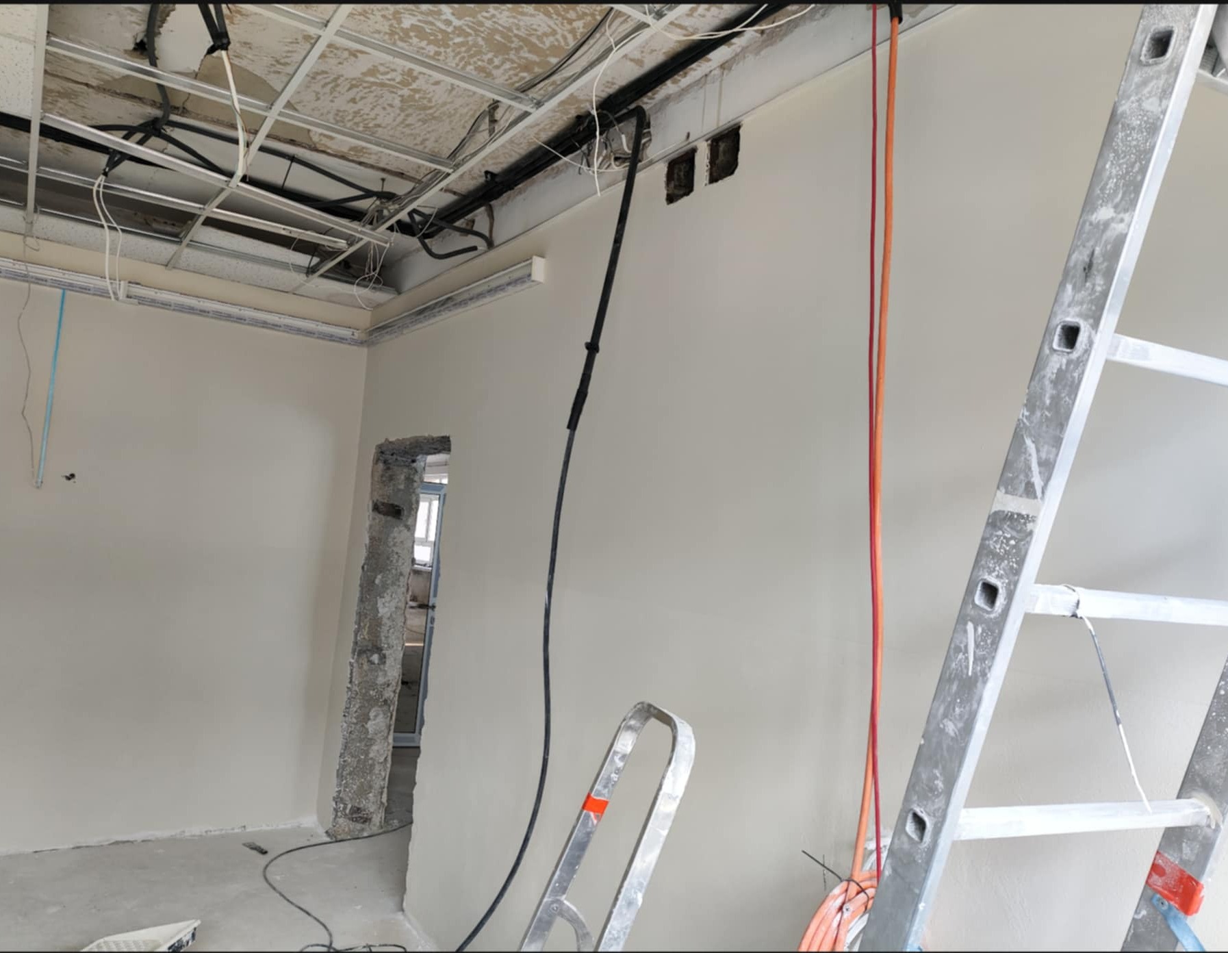 Wnętrze w trakcie prac wykończeniowych: świeżo wyszpachlowana ściana, widoczne kable, drabina aluminiowa, sufit z instalacjami. Otwór drzwiowy w stanie surowym.