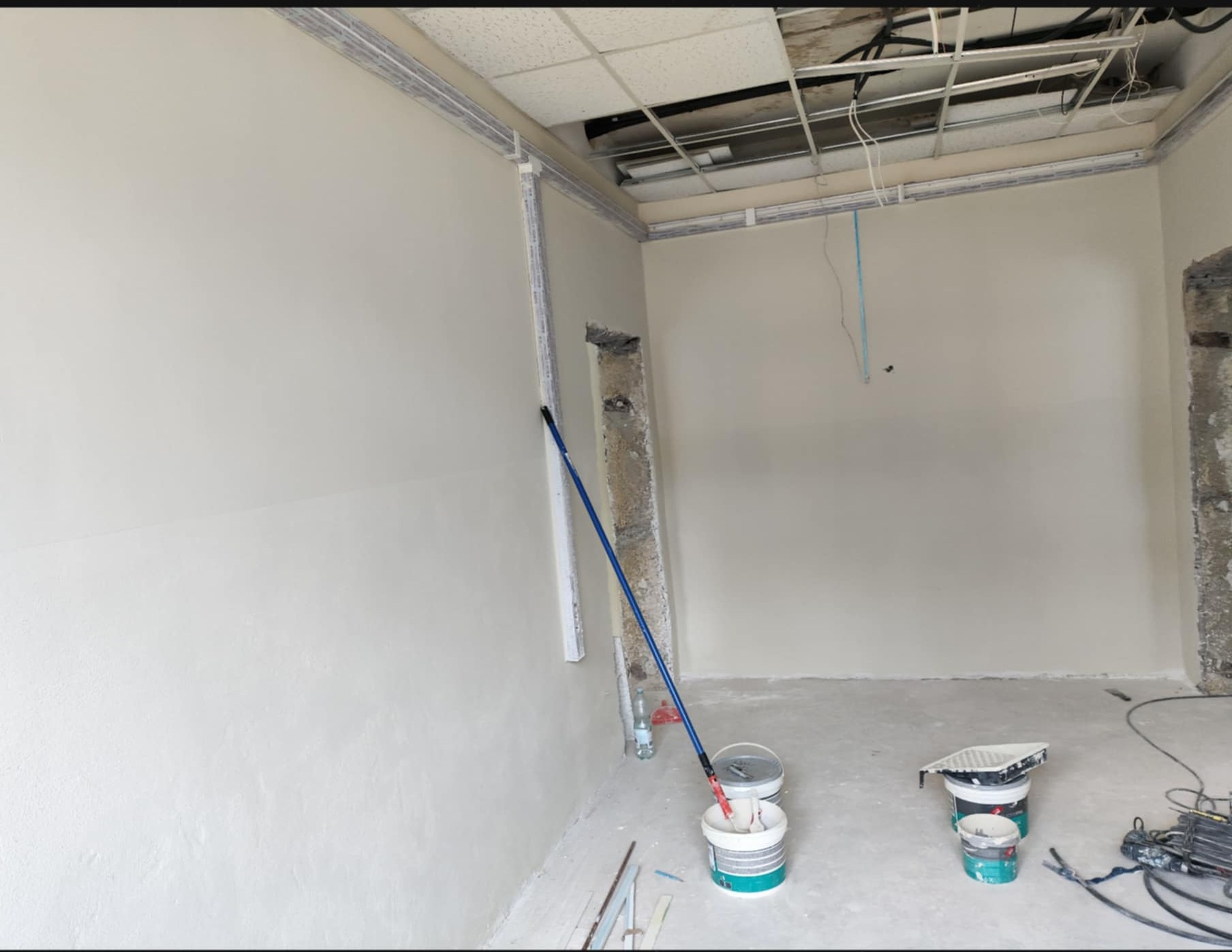 Wnętrze pokoju w trakcie prac wykończeniowych; świeżo pomalowane ściany na biało, widoczne narzędzia malarskie i puszki z farbą. Sufit z instalacją.