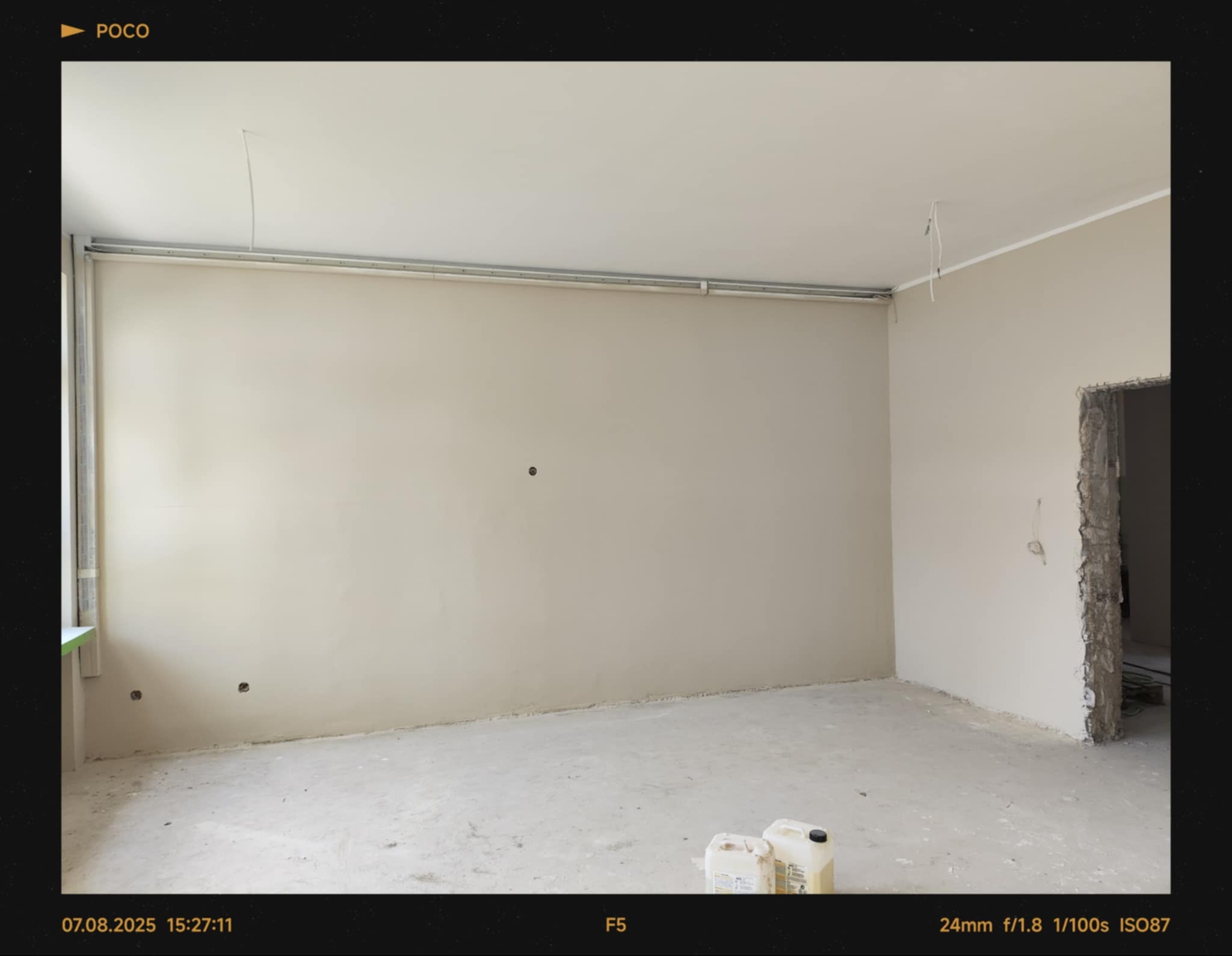 Wnętrze pokoju po szpachlowaniu ścian, przygotowane do malowania. Widoczne gładkie ściany, instalacje pod sufitem i kanistry z farbą na podłodze.