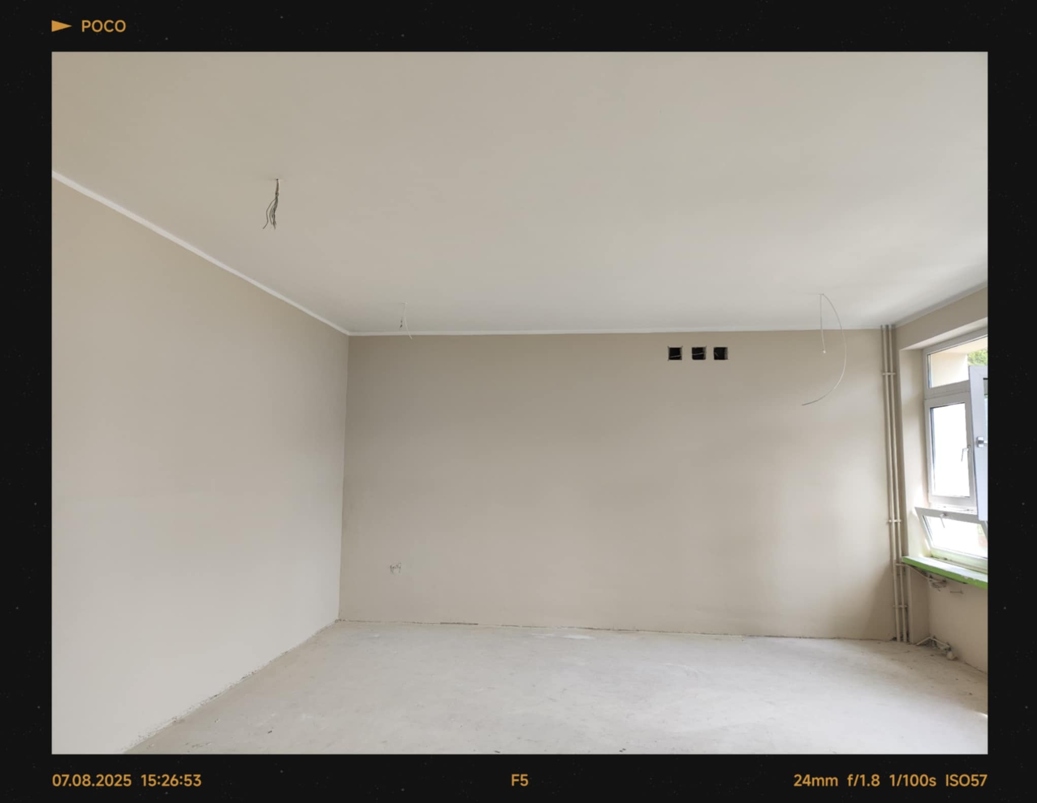 Wnętrze pokoju po remoncie. Ściany i sufit pomalowane na jasny, beżowy kolor. Widoczne wystające kable, listwy przypodłogowe i okno z parapetem.