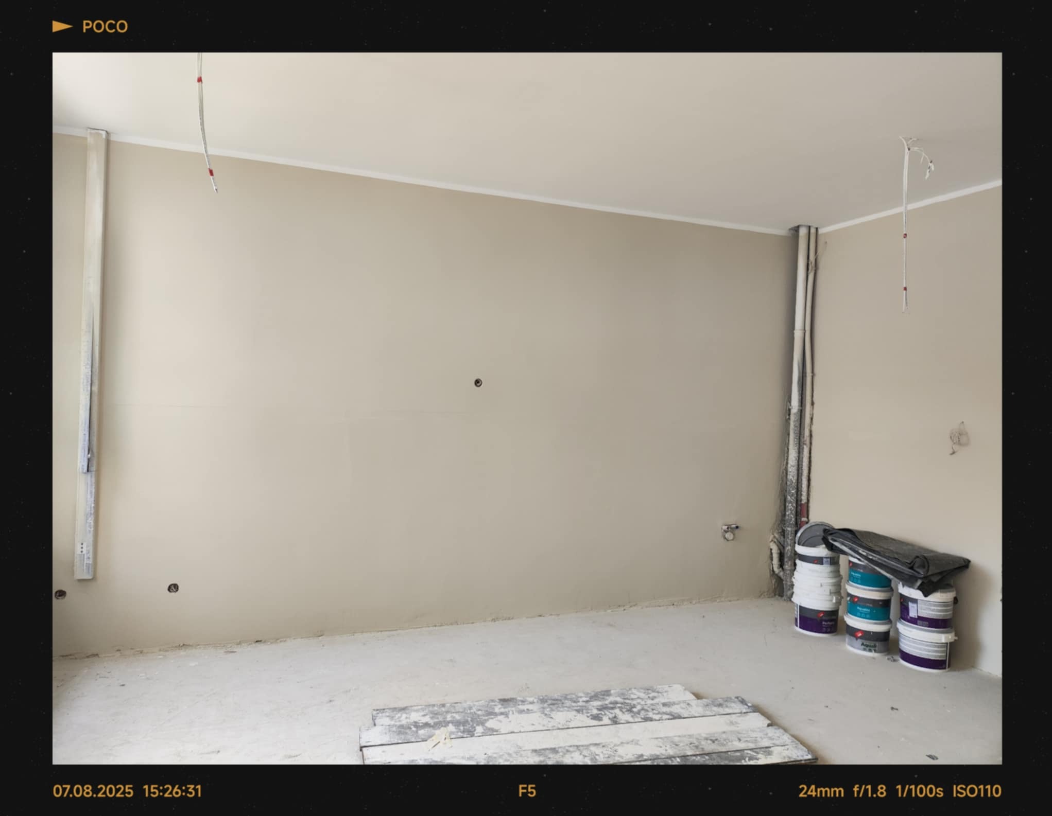 Pokój w trakcie malowania: ściany w beżowym odcieniu, widoczne puszki z farbą, przygotowane do dalszych prac wykończeniowych. Minimalizm i stonowana kolorystyka.