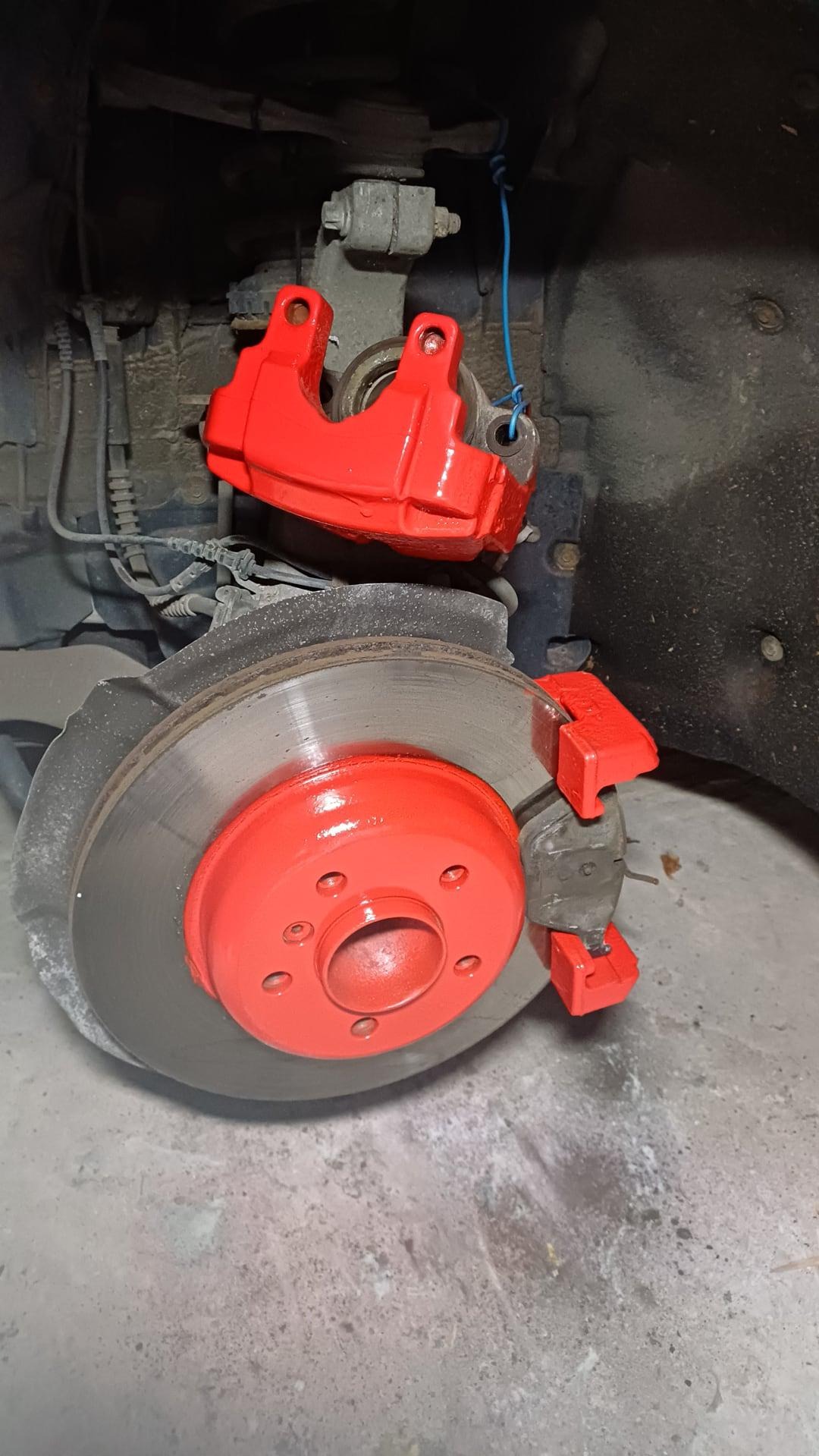 Czerwone zaciski hamulcowe po tuningu, widoczne na tle tarczy hamulcowej i zawieszenia. Detal z niebieskim przewodem dodaje unikalności ujęciu.