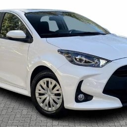 Royal Fleet sp zoo - Biały samochód Toyota Yaris, widok z przodu na szarym brukowanym podłożu, z logo producenta na masce.