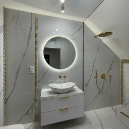 Welldonebathrooms.com - Wykończona łazienka na poddaszu: białe płytki imitujące marmur z złotymi akcentami, okrągłe lustro z podświetleniem LED, biała szafka pod umywalką i złota armatura.