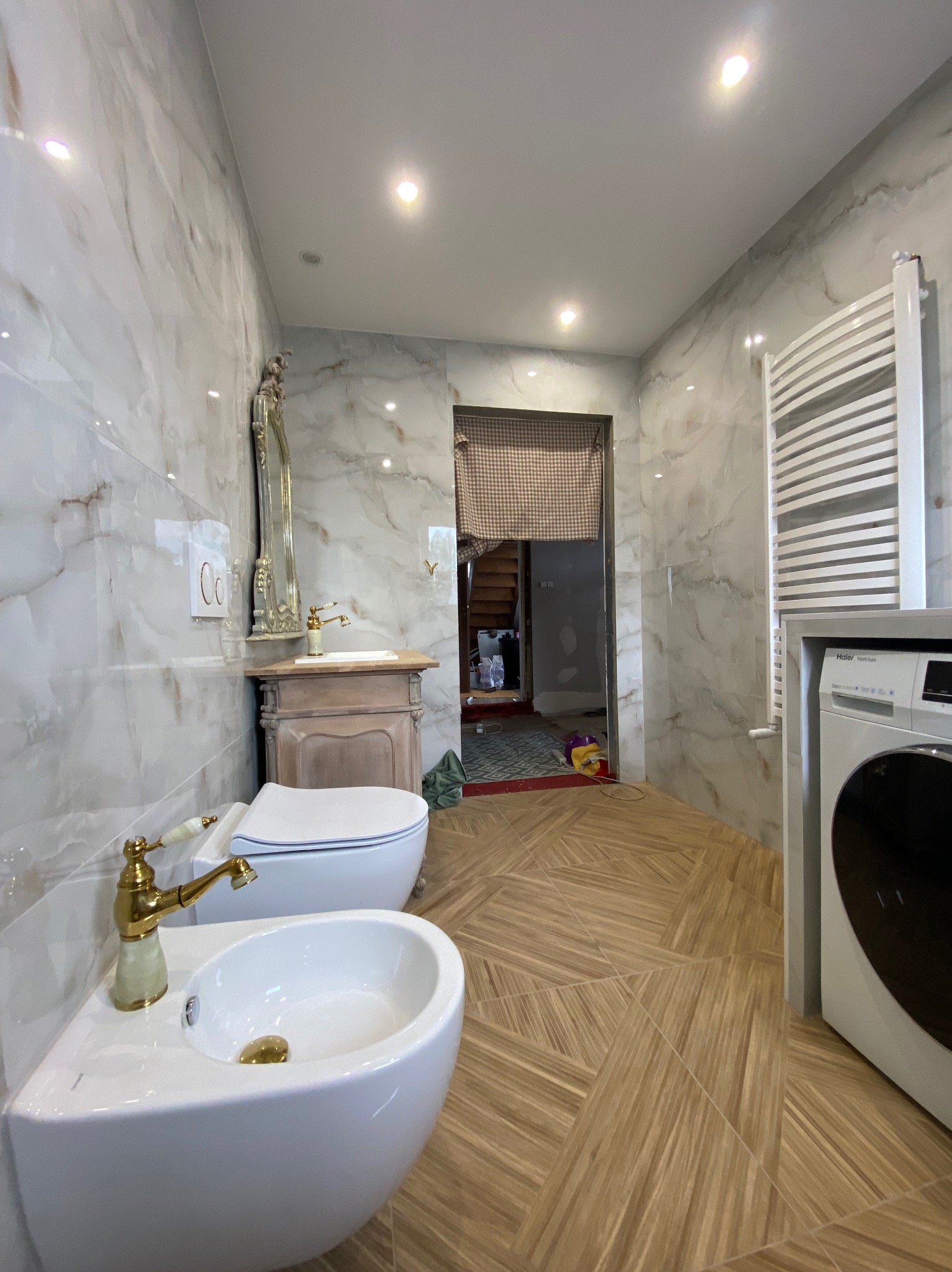 Jasna łazienka z marmurowymi płytkami, drewnopodobną podłogą, toaletą z bidetem i złotą armaturą, oraz pralką i grzejnikiem drabinkowym. W tle widoczne niedokończone prace remontowe.