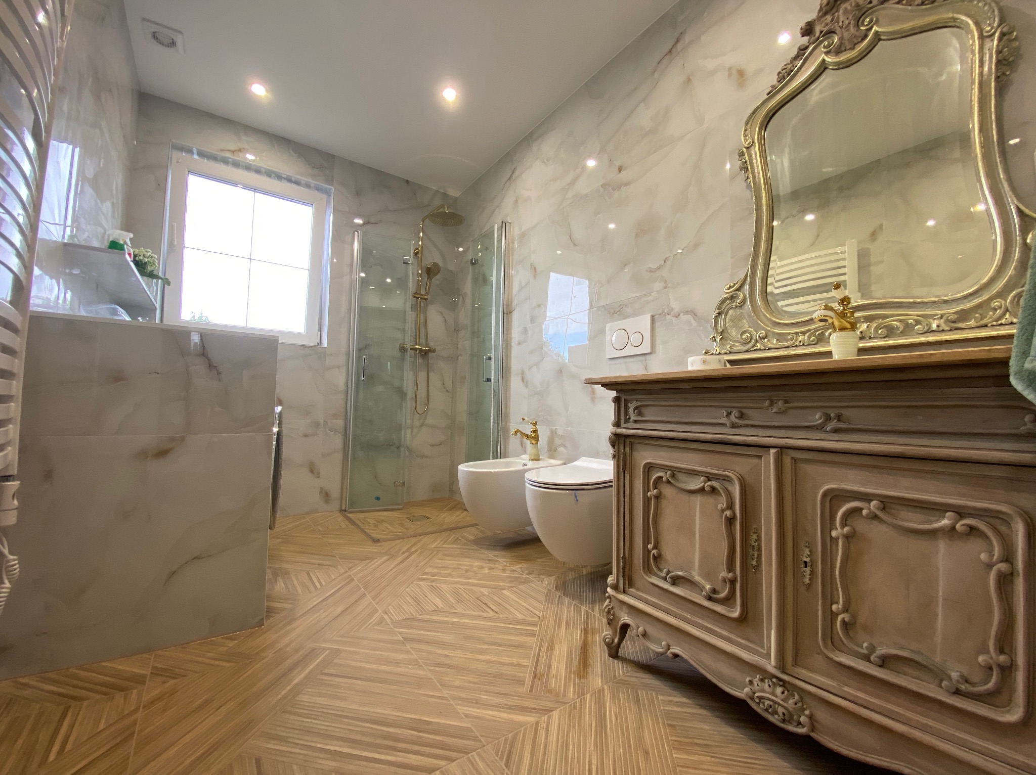 Elegancka łazienka z marmurowymi płytkami, drewnianą podłogą w geometryczny wzór i ozdobnym lustrem nad stylizowaną komodą.