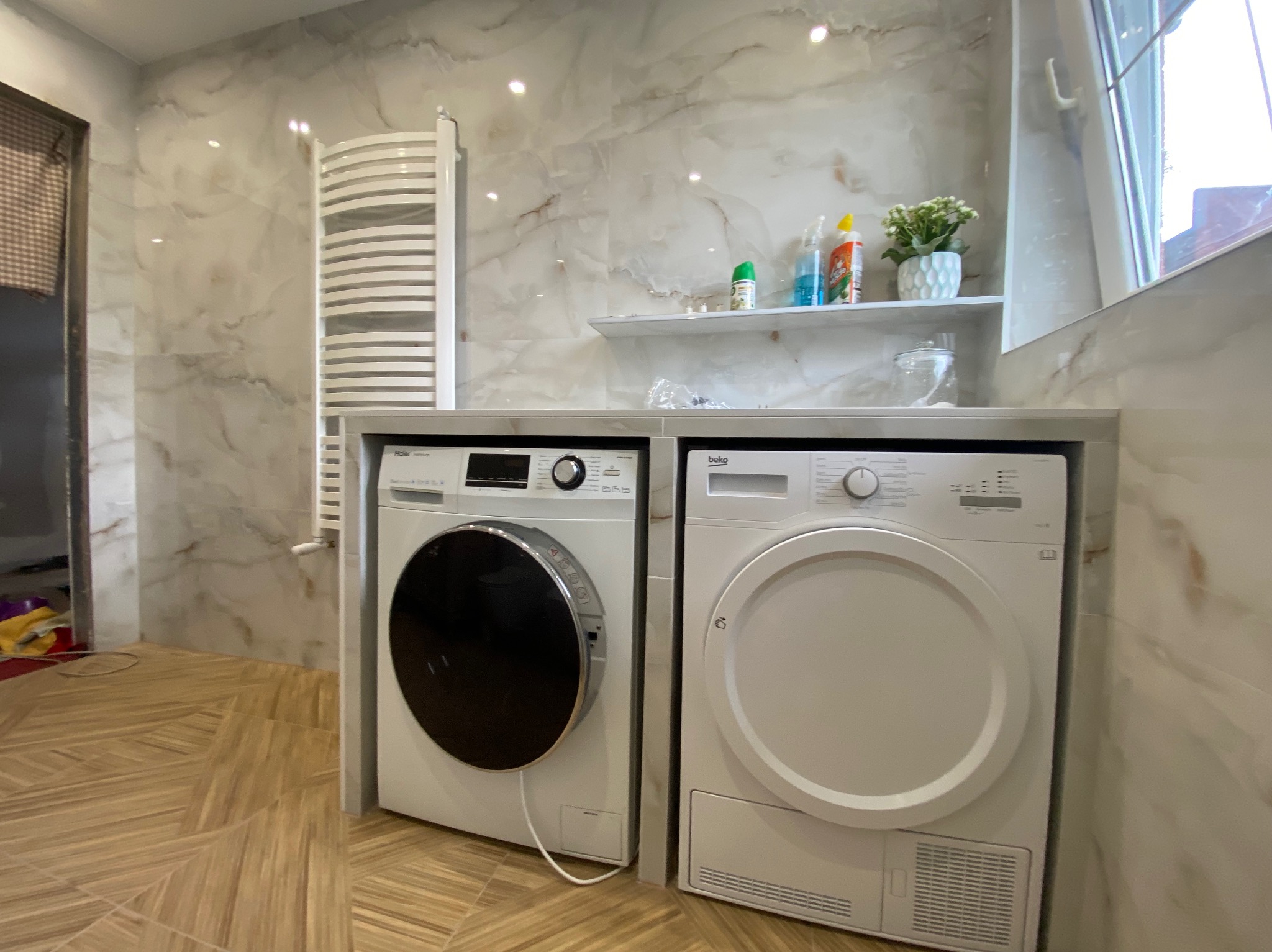 Wykończona łazienka z marmurowymi płytkami, grzejnikiem drabinkowym i zabudowaną pralką oraz suszarką Beko pod blatem z półką, na której stoją środki czystości i doniczka z kwiatami.
