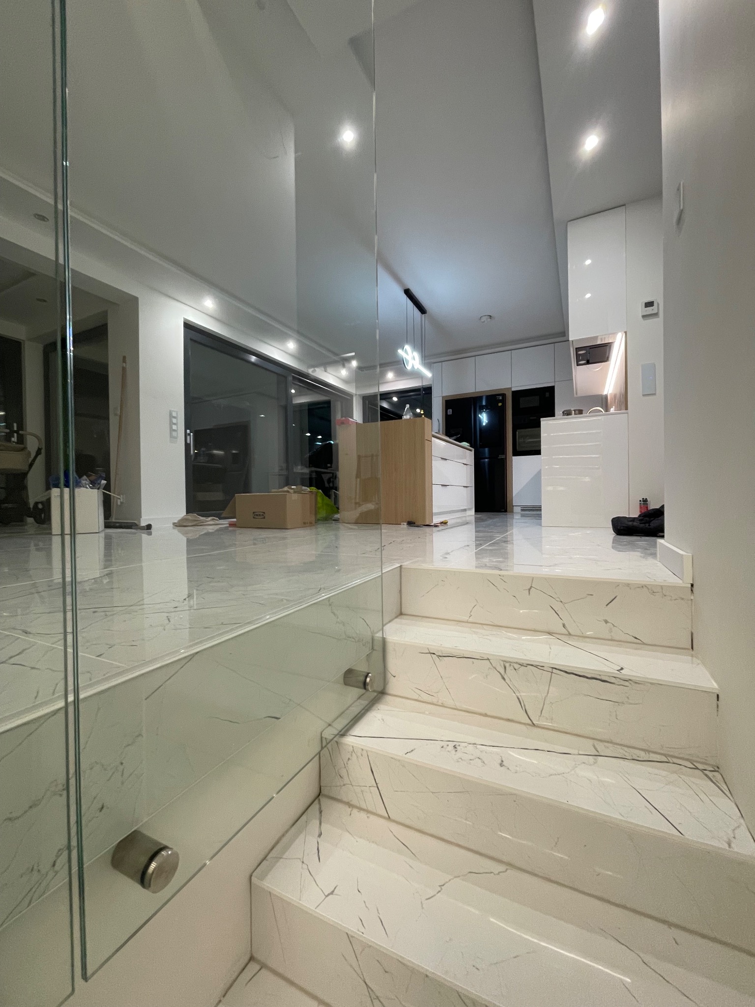 Białe marmurowe schody z szklaną balustradą prowadzą do nowoczesnej kuchni z białymi szafkami i błyszczącą podłogą. Widoczne kartony sugerują niedawne wykończenie.