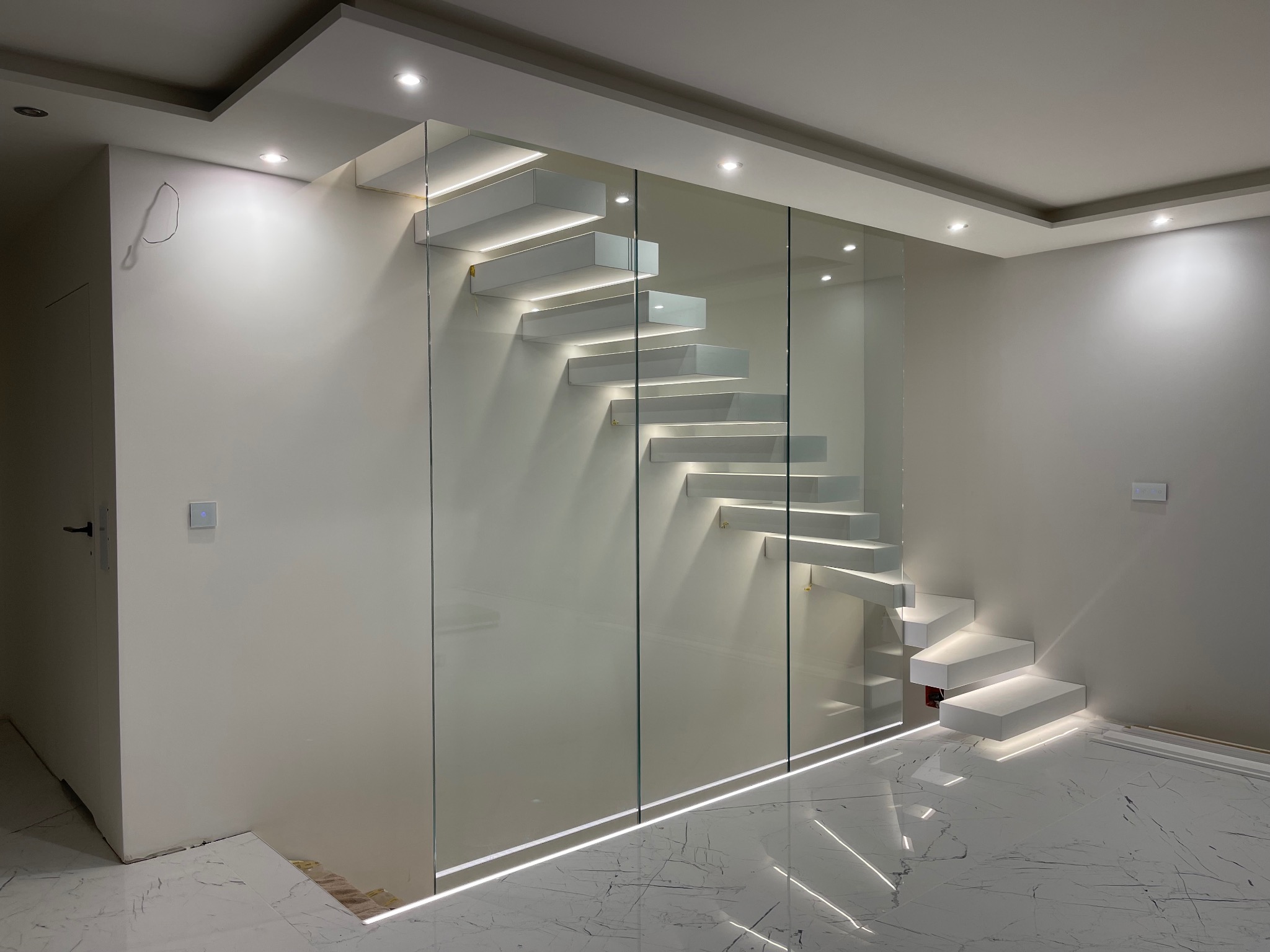 Nowoczesne, podświetlane schody wiszące z białymi stopniami i szklaną balustradą, oświetlenie LED w podłodze, marmurowa posadzka, minimalistyczny design.