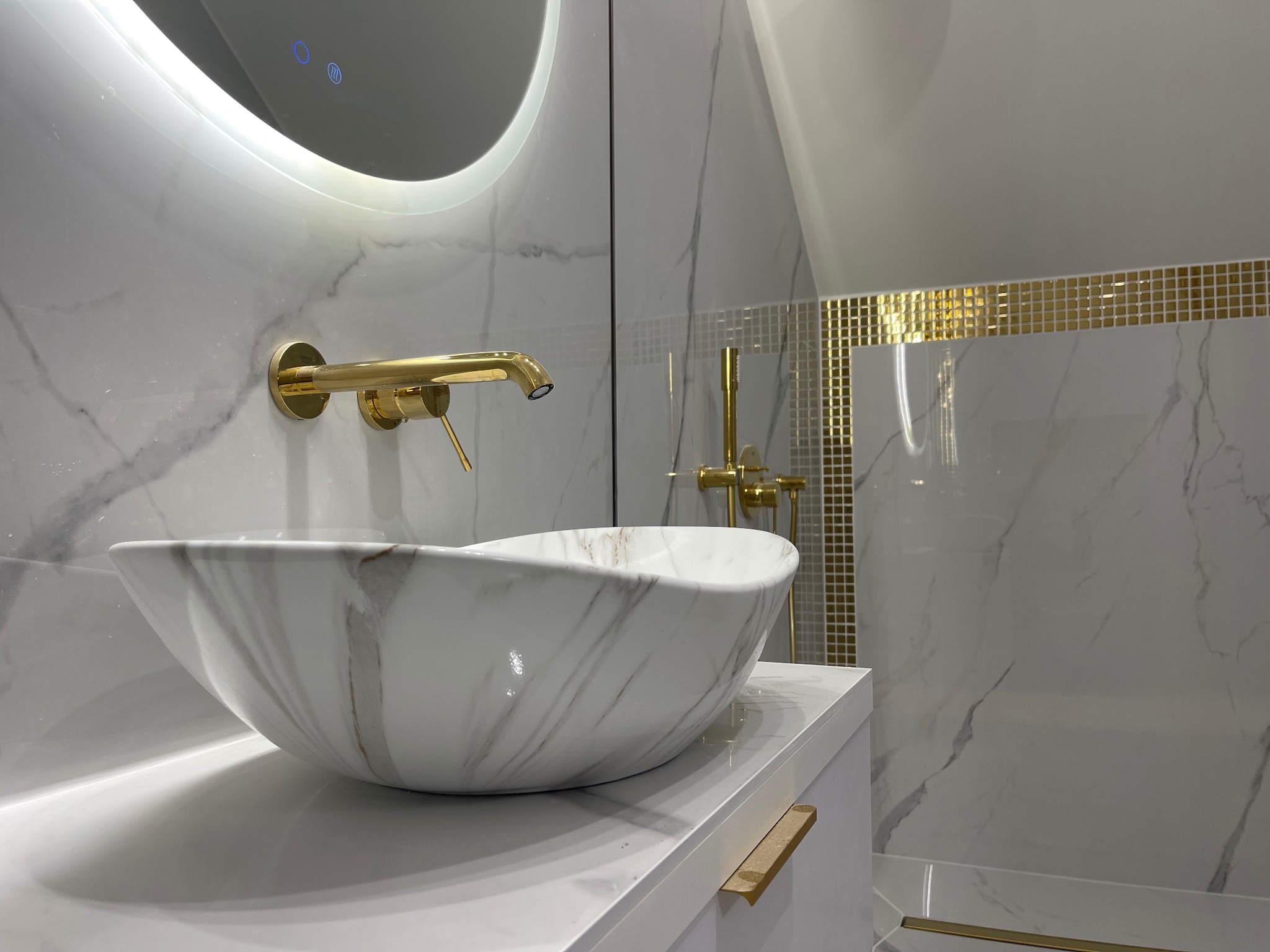Elegancka łazienka: biała umywalka marmurkowa, złota armatura i mozaika. Podświetlane lustro dodaje blasku. Nowoczesny design z dbałością o detale.