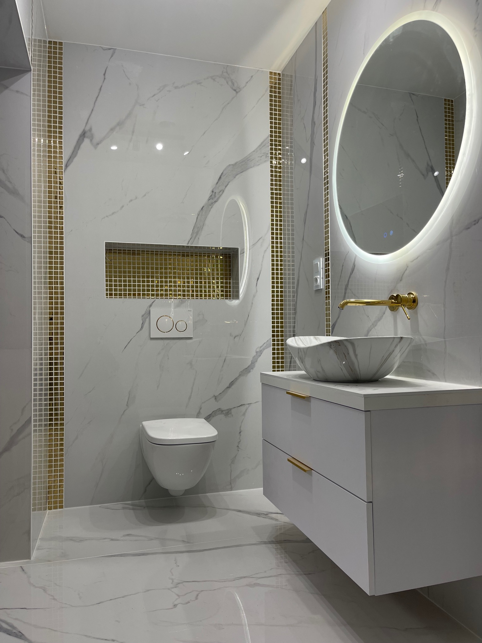 Elegancka łazienka w bieli i złocie: marmurowe płytki, owalne lustro z podświetleniem LED, wisząca toaleta i umywalka nablatowa z armaturą w kolorze złota.