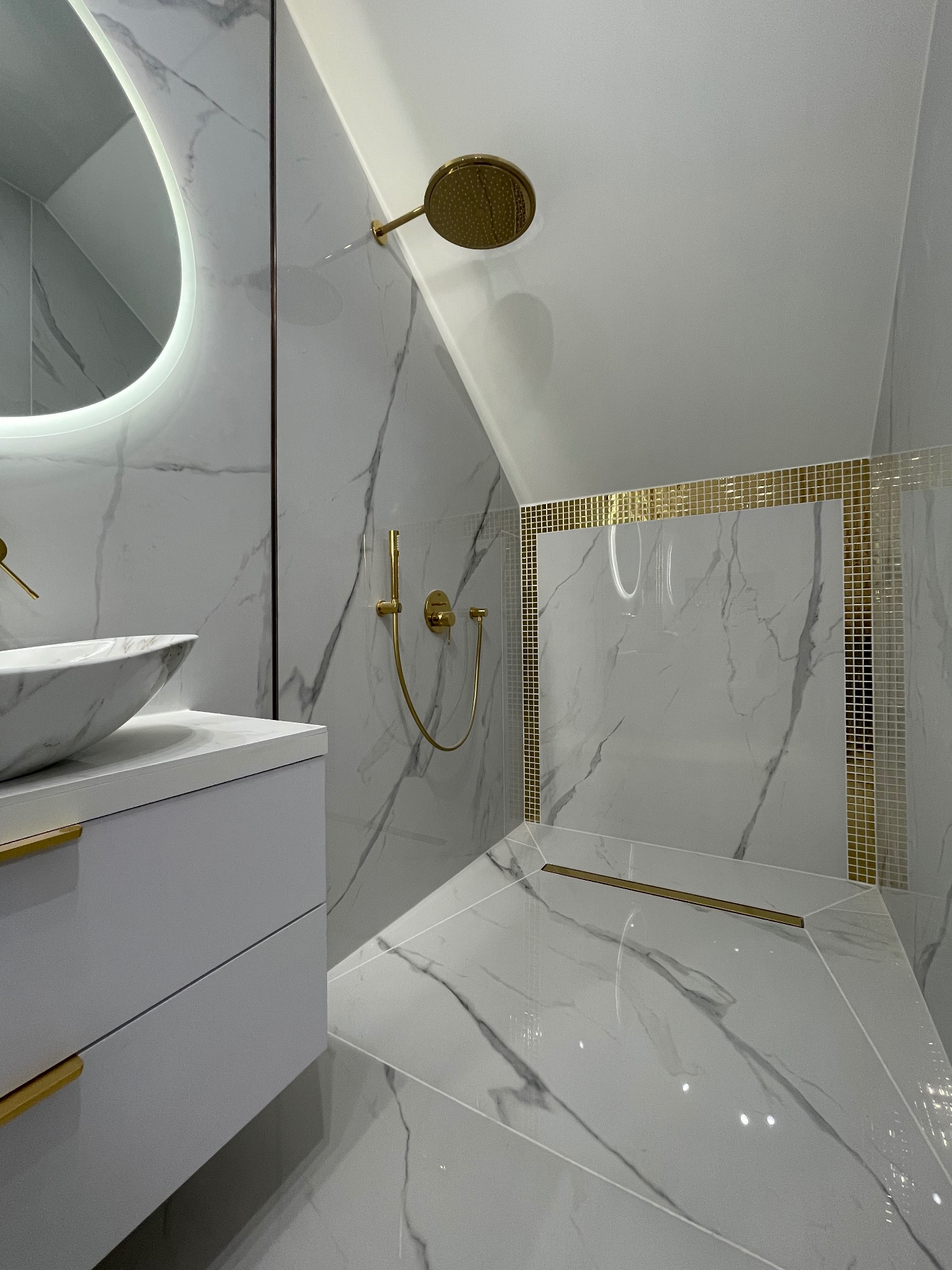 Elegancka łazienka na poddaszu: białe marmurowe płytki, złote akcenty w armaturze i mozaice, owalne lustro z podświetleniem, nowoczesna umywalka nablatowa.