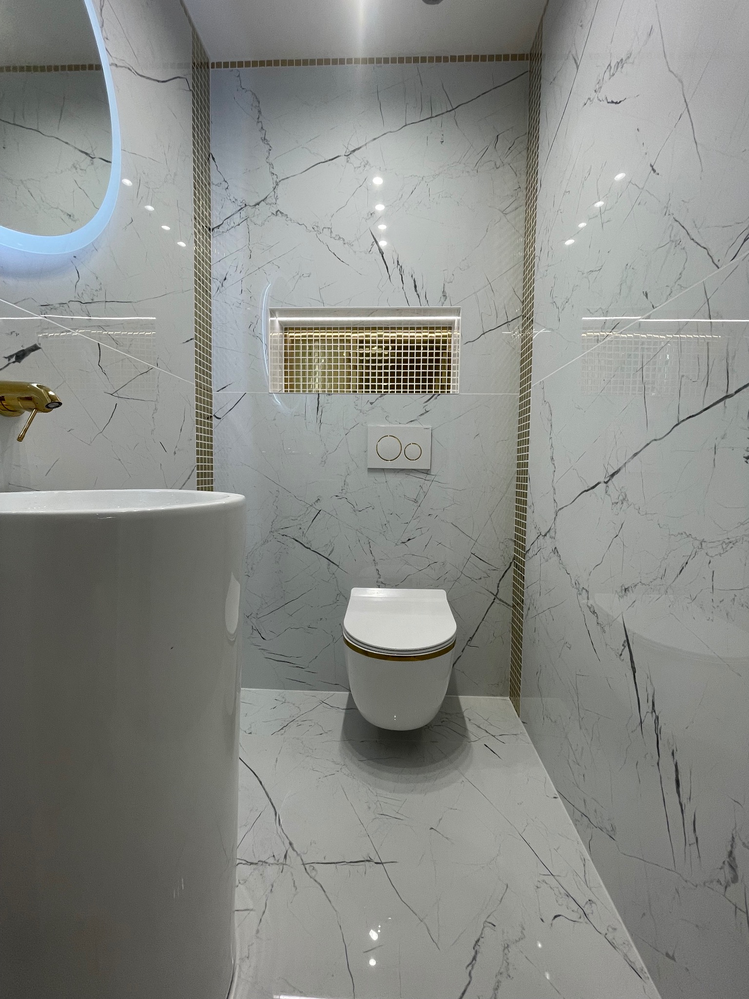 Elegancka toaleta w bieli i złocie: marmurowe płytki, podświetlane lustro i designerska armatura tworzą luksusową przestrzeń. Minimalistyczny styl z dbałością o detale.