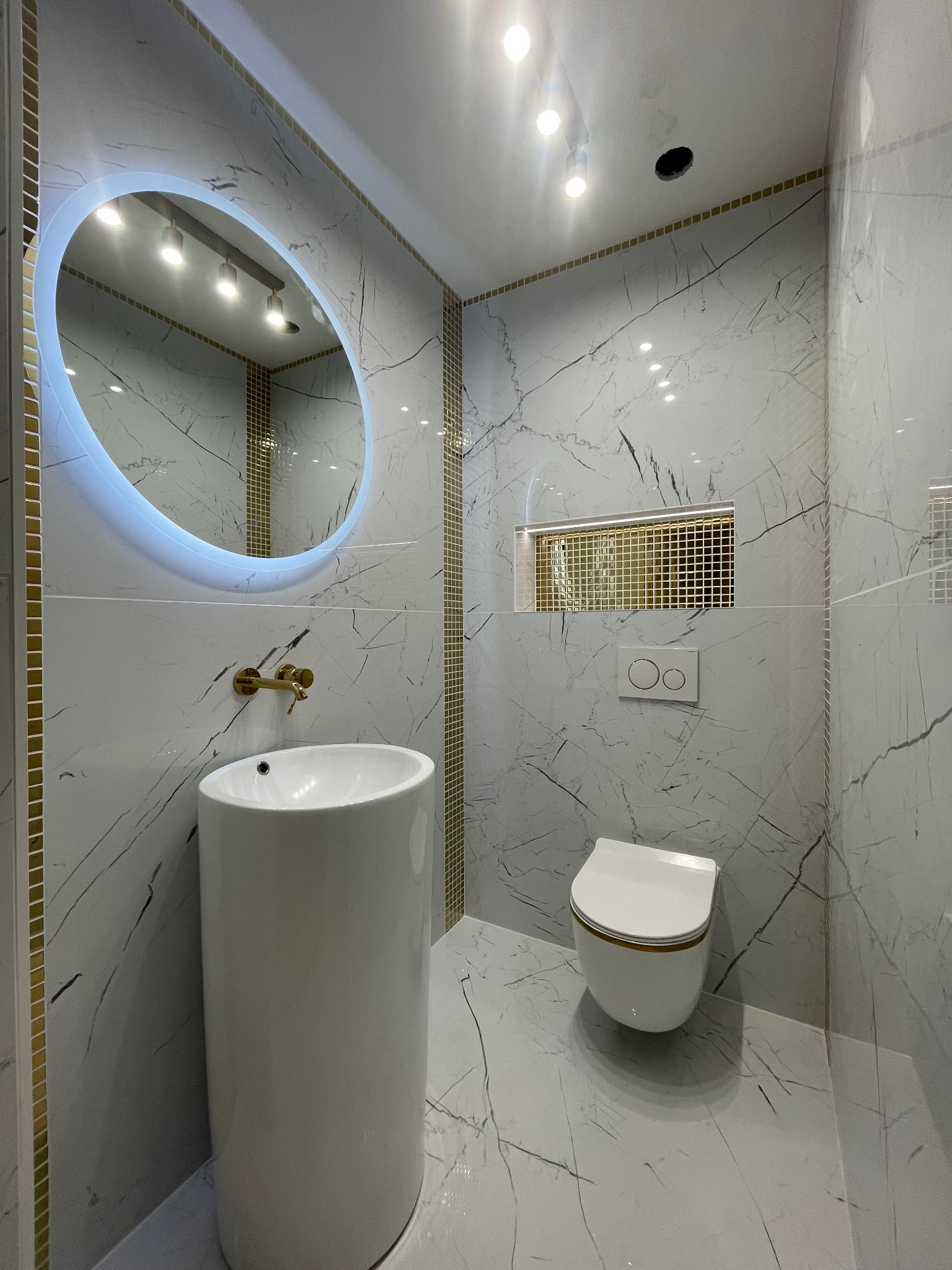 Elegancka łazienka z białymi płytkami imitującymi marmur, okrągłym lustrem z podświetleniem LED i złotymi akcentami. Umywalka kolumnowa i podwieszana toaleta.