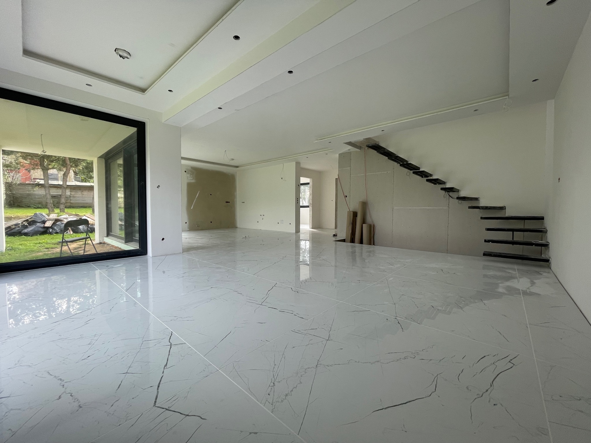 Jasne, marmurowe płytki podłogowe w przestronnym wnętrzu z surowymi schodami i dużym oknem. Widoczne przygotowania do dalszych prac wykończeniowych.