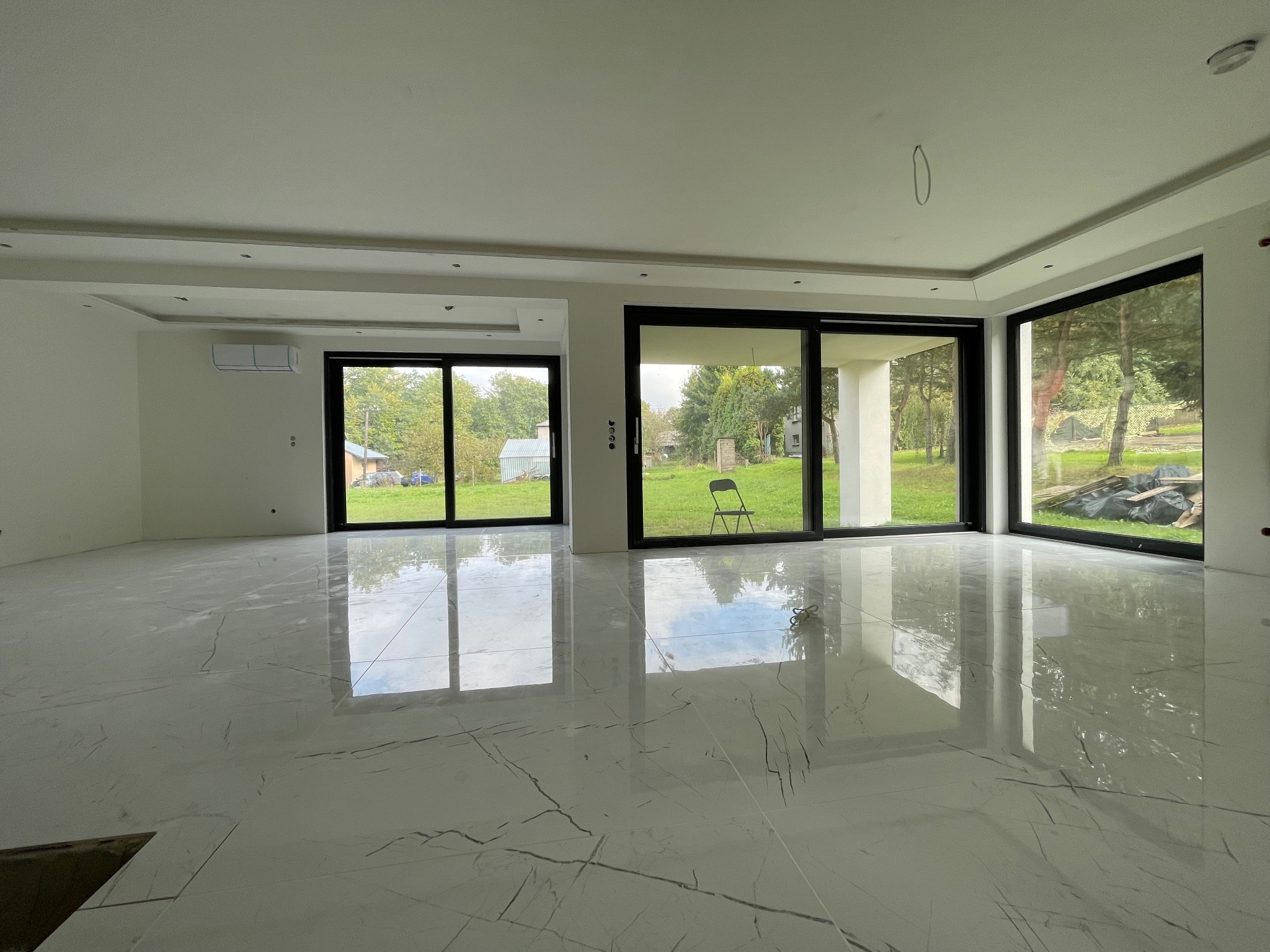 Błyszcząca, biała podłoga z marmurowym wzorem w przestronnym, jasnym wnętrzu z dużymi oknami. Widoczny fragment sufitu z oświetleniem.