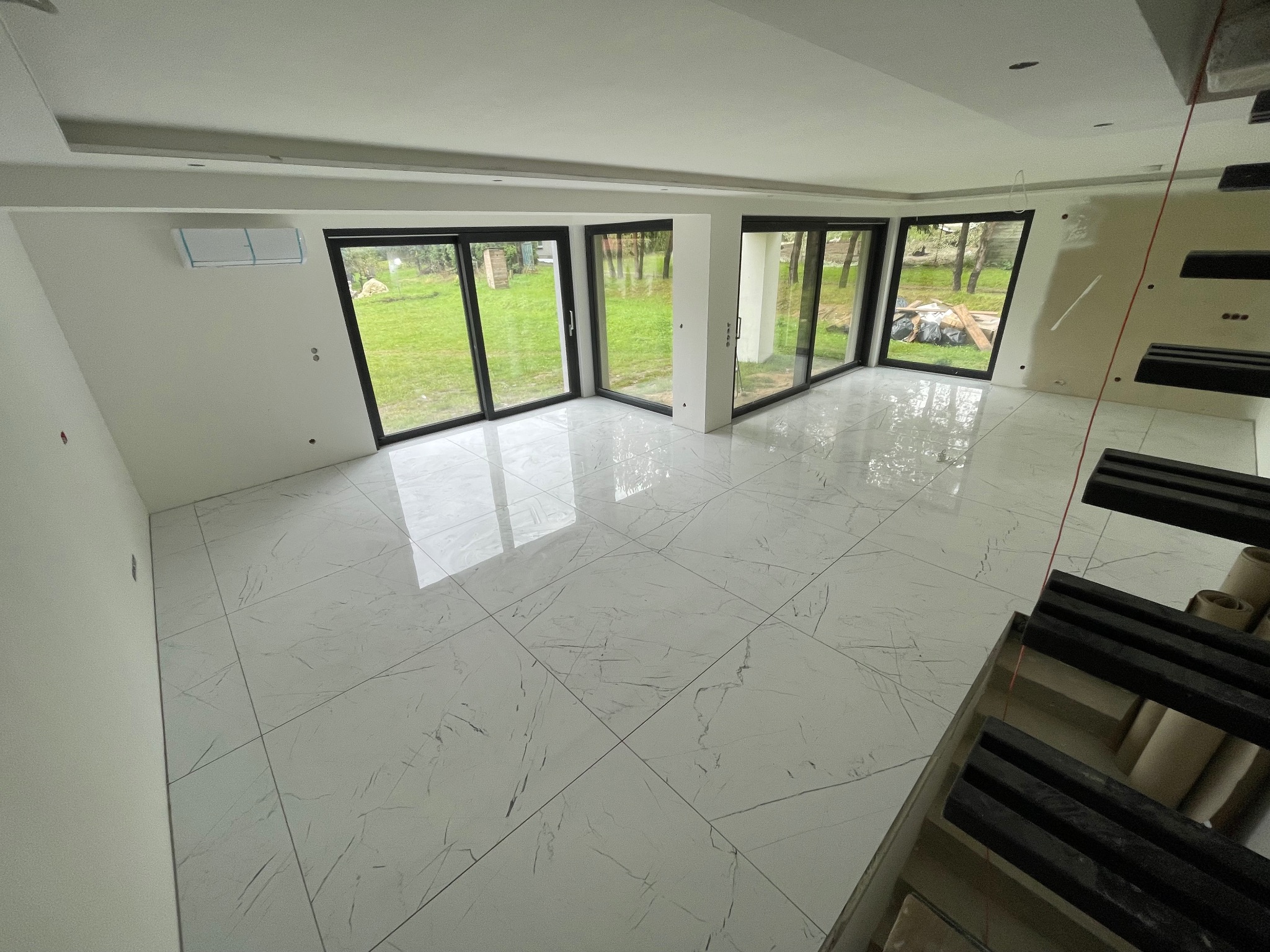 Jasne, marmurowe płytki podłogowe w przestronnym wnętrzu z dużymi oknami. Widoczne niedokończone schody i klimatyzator. Minimalistyczny design.