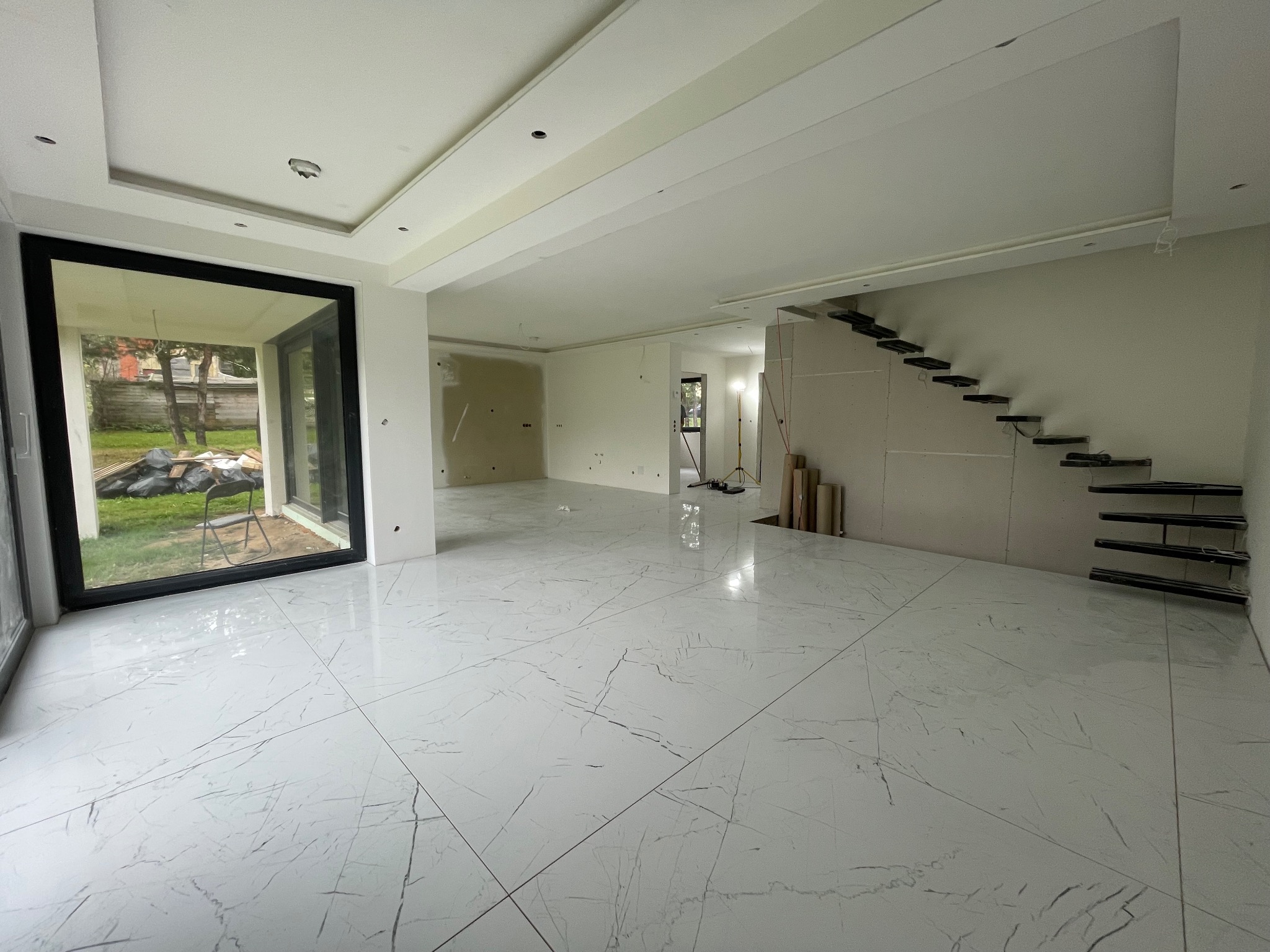 Jasne, marmurowe płytki podłogowe w przestronnym wnętrzu z dużym oknem i schodami. Widoczne elementy wykończeniowe sufitu i ścian. Minimalistyczny design.