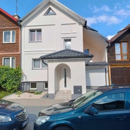 Lukbud - Dwupiętrowy dom z białym tynkiem, szarym dachem, podjazdem z kostki brukowej i dwoma zaparkowanymi samochodami na pierwszym planie.