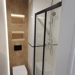 Lukbud - Nowoczesna łazienka z kabiną prysznicową z czarnymi ramami, podświetlaną ścianą z drewnopodobnych płytek ułożonych w jodełkę i wiszącą toaletą.