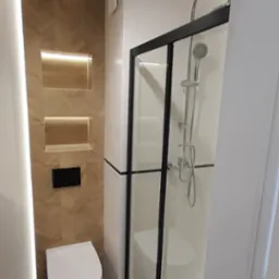 Nowoczesna łazienka z kabiną prysznicową z czarnymi ramami, podświetlaną ścianą z drewnopodobnych płytek ułożonych w jodełkę i wiszącą toaletą.