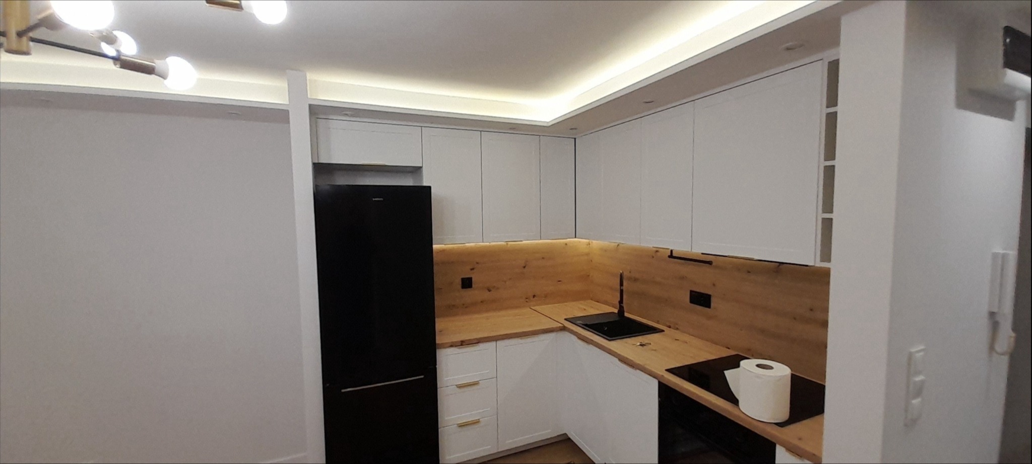 Nowoczesna kuchnia z białymi szafkami, drewnianym blatem i czarną lodówką, oświetlona listwą LED pod szafkami wiszącymi.