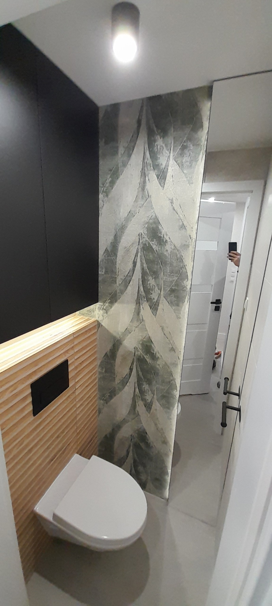 Nowoczesna łazienka z wiszącą toaletą, drewnianą okładziną ścienną z czarnym przyciskiem spłukującym i tapetą w liście w odcieniach szarości, odbicie osoby robiącej zdjęcie w lustrze.