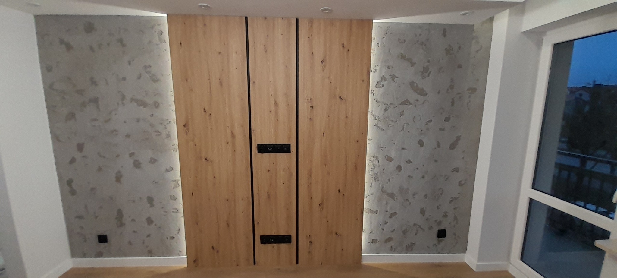 Wykończona ściana z drewnianymi panelami z czarnymi wstawkami i gniazdkami elektrycznymi, obok ściana z dekoracyjnym tynkiem imitującym kamień i białym oknem z widokiem na zewnątrz.