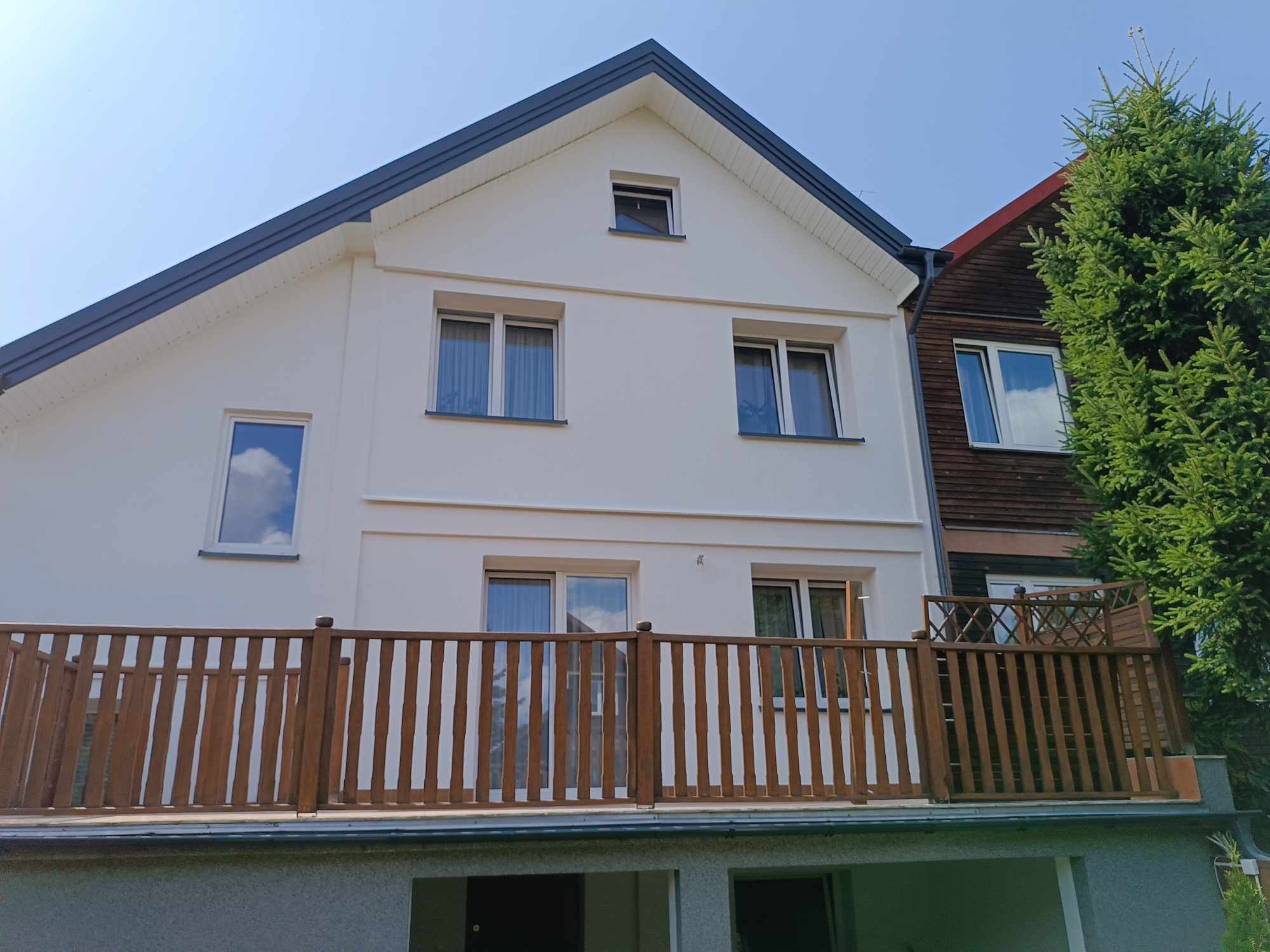 Odnowiona, dwupiętrowa fasada domu z białym tynkiem, grafitowym dachem i drewnianą balustradą balkonu na pierwszym piętrze. Widoczne okna z białymi ramami i odbijające się niebo.