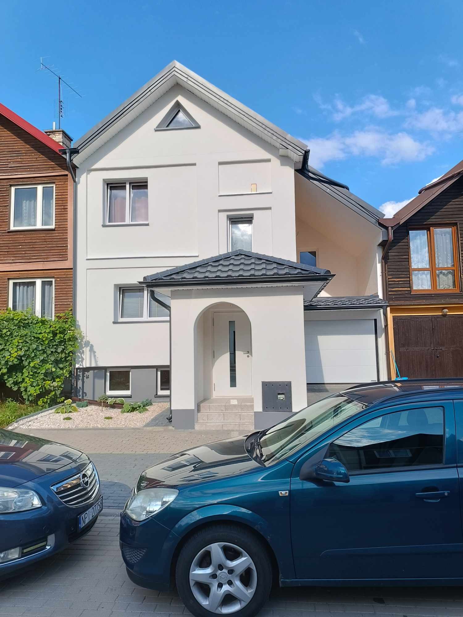 Dwupiętrowy dom z białym tynkiem, szarym dachem, podjazdem z kostki brukowej i dwoma zaparkowanymi samochodami na pierwszym planie.