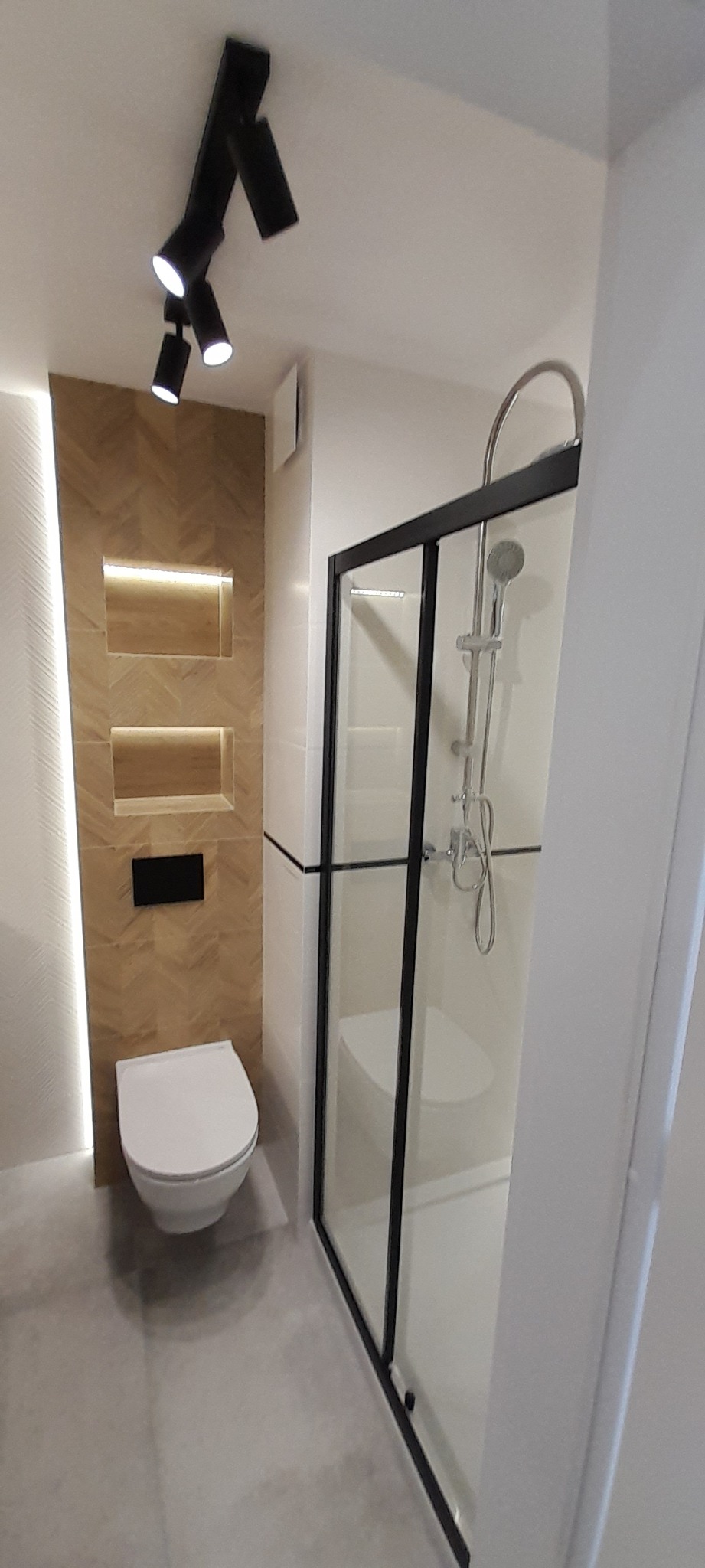Nowoczesna łazienka z kabiną prysznicową z czarnymi ramami, podświetlaną ścianą z drewnopodobnych płytek ułożonych w jodełkę i wiszącą toaletą.