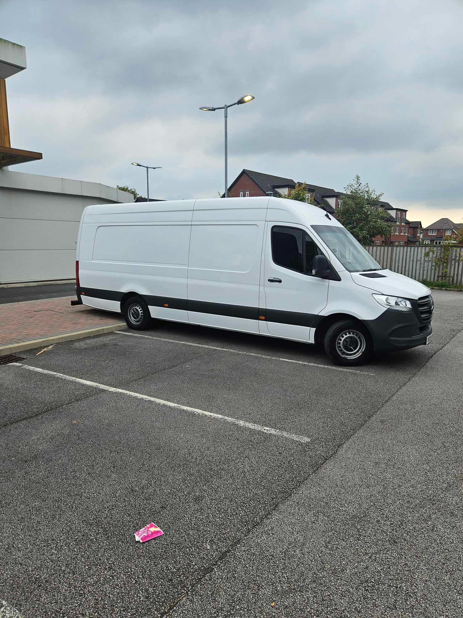 Biały samochód dostawczy zaparkowany na parkingu, w tle budynki mieszkalne i pochmurne niebo; na ziemi leży różowe opakowanie.