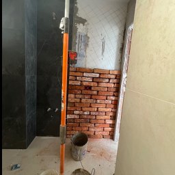 Aleksander Smoleń VentiPRO - Remont łazienki: fragment ściany z cegieł, obok czarne płytki imitujące marmur. Widoczne narzędzia i wiaderka. Etap prac przed wykończeniem.