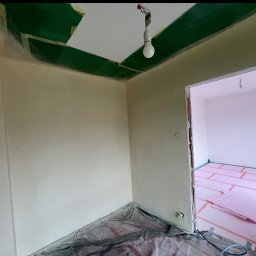 Aleksander Smoleń VentiPRO - Świeżo pomalowane ściany w pokoju, zabezpieczone folią i taśmą malarską, z widocznym ociepleniem podłogi w sąsiednim pomieszczeniu. Żarówka zwisa z sufitu.