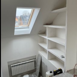 Aleksander Smoleń VentiPRO - Białe, wnękowe półki w skosie poddasza, widok na okno dachowe i grzejnik. Ślady prac wykończeniowych na podłodze. Detal wnętrza po remoncie.