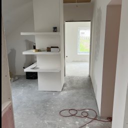 Aleksander Smoleń VentiPRO - Wnętrze w trakcie wykończenia: surowy beton na podłodze, białe ściany po gładzi, regał z półkami. Widoczny kabel na podłodze i okno w oddali.