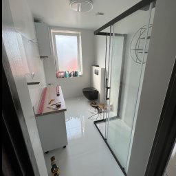 Aleksander Smoleń VentiPRO - Biała łazienka w trakcie wykańczania: kabina prysznicowa z czarnymi ramami, umywalka z blatem, czarna toaleta. Widoczne narzędzia i poziomice.